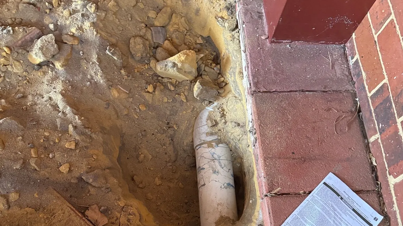 Hand-dug hole in yellow sand exposing a PVC drain pipe beside red brick paving at a Craigie home